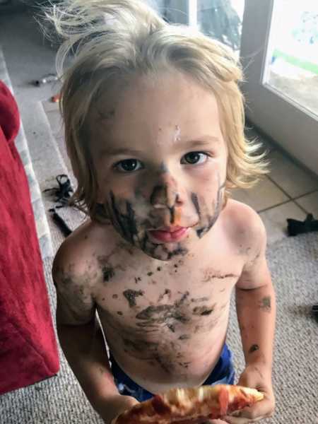 Young boy looks up covered in dirt while holding a piece of pizza