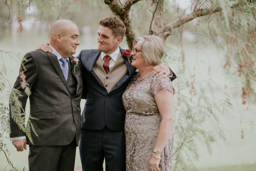 Groom stands arm in arm with mother and father who has cancer