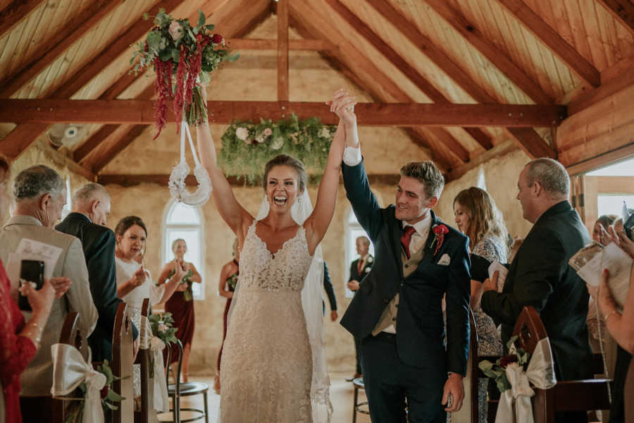 Bride and groom whose father has cancer holds arms up in air of aisle after being married