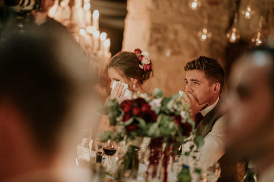Groom whose father has cancer sits choked up at wedding reception next to bride who wipes his tears