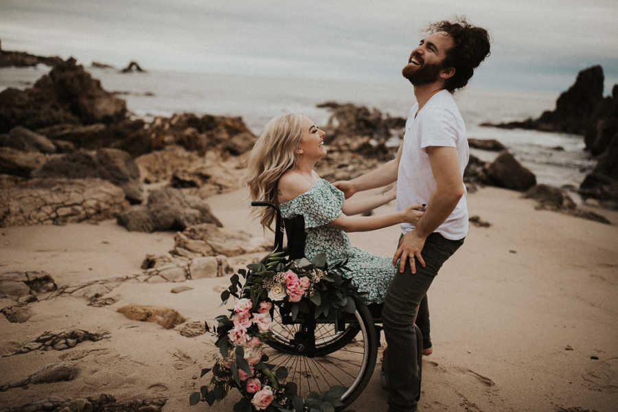 Woman in wheelchair laughs as she holds on to boyfriends waist on beach
