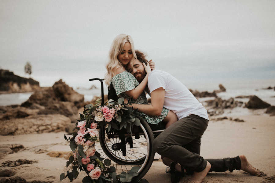 Boyfriend kneels and leans over to hug girlfriend in wheelchair on beach