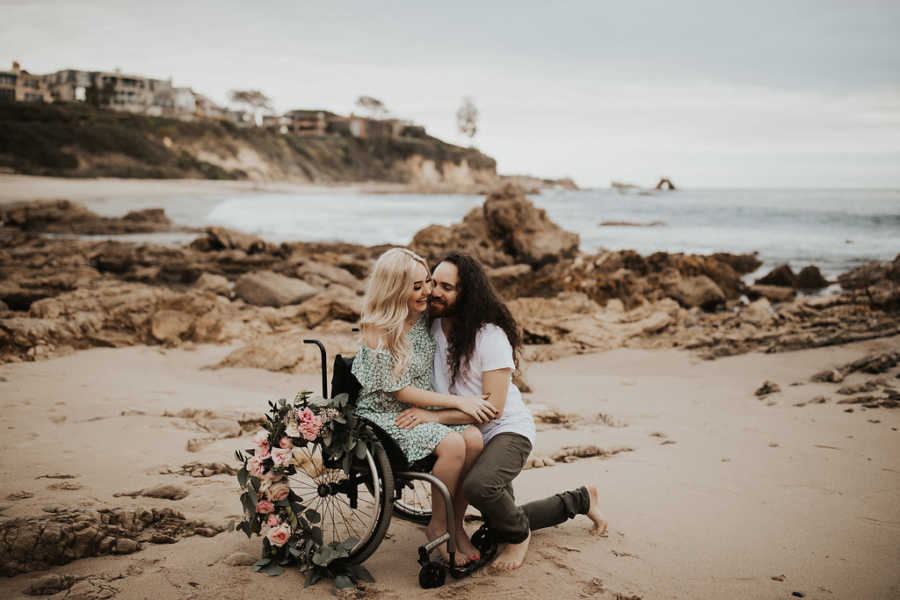 Woman in wheelchair smiles as boyfriend kneels next to her hugging her