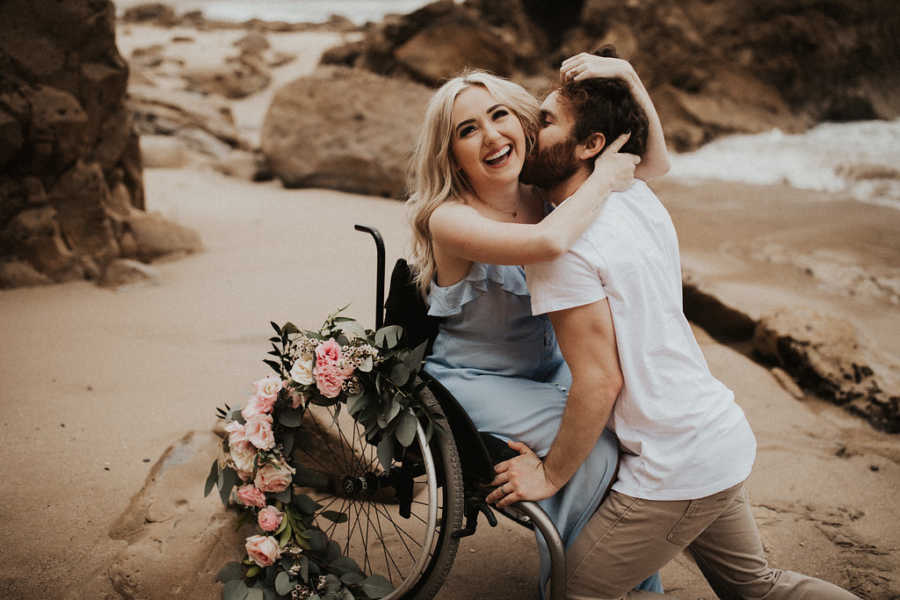 Woman with Spinal Muscular Atrophy sits in wheelchair on beach while boyfriend kisses her
