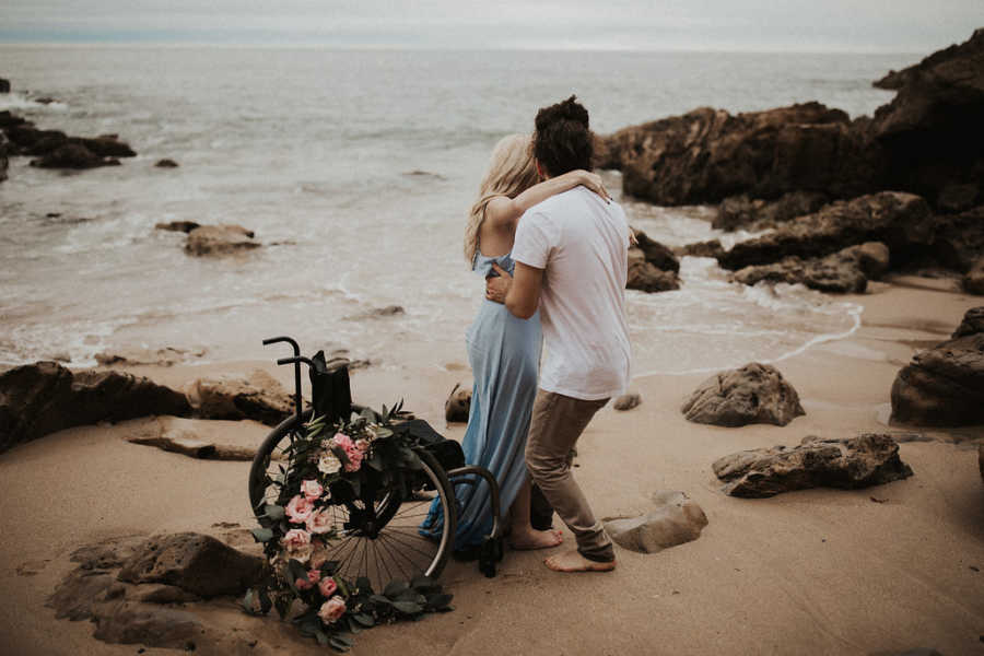 Boyfriend holds girlfriend with wheelchair in his arms as they stand on beach