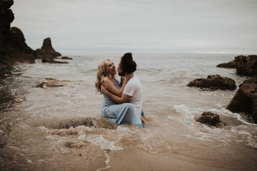 Girlfriend with Spinal Muscular Atrophy sits on lap of boyfriend on beach with tide covering them