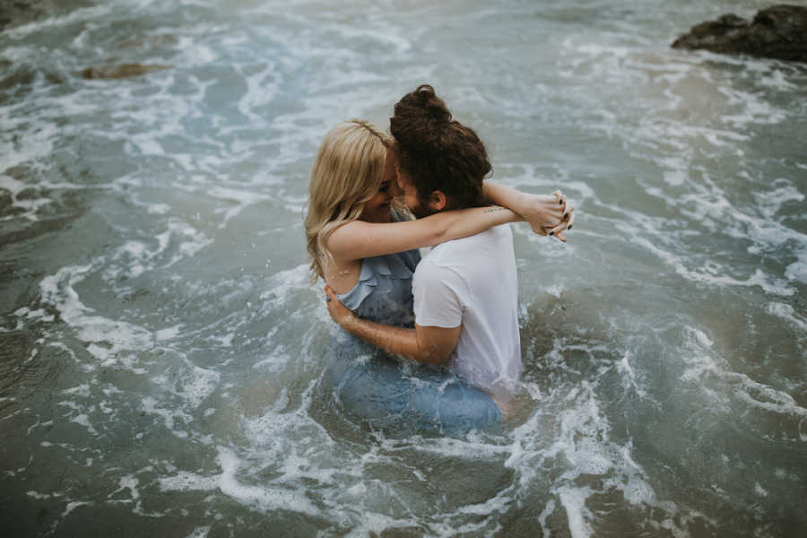 Girlfriend with Spinal Muscular Atrophy sits on boyfriends lap with arms wrapped around him in ocean