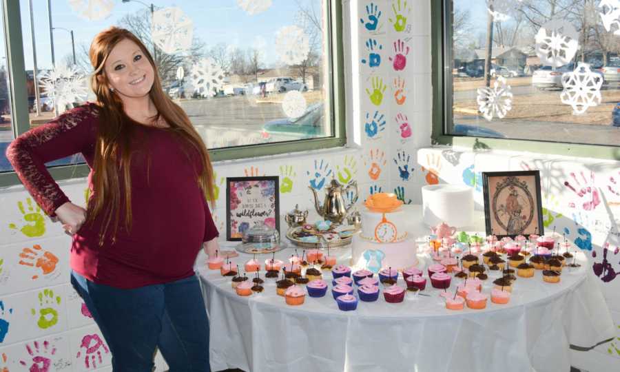 Pregnant woman stands beside table at baby shower with pink cupcakes and hand prints all over wall