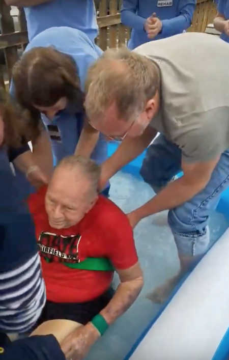 Elderly man who had stroke sitting in inflatable pool being baptized with son at his side