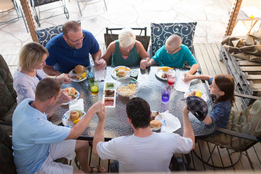 Family with three dads, two moms, and two children hold hands and pray at table outdside