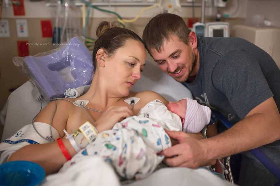 Mother who just had c-section sits in hospital bed holding newborn while husband leans over to look at baby