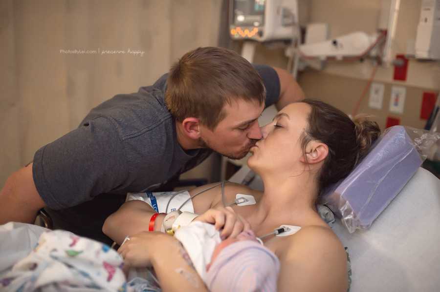 Husband leans over to kiss wife in hospital bed who holds newborn after c-section
