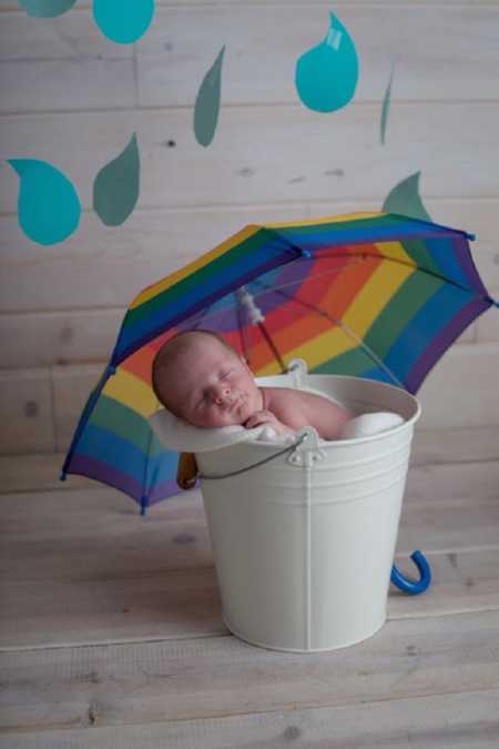 Newborn sitting in bucket with rainbow umbrella above it