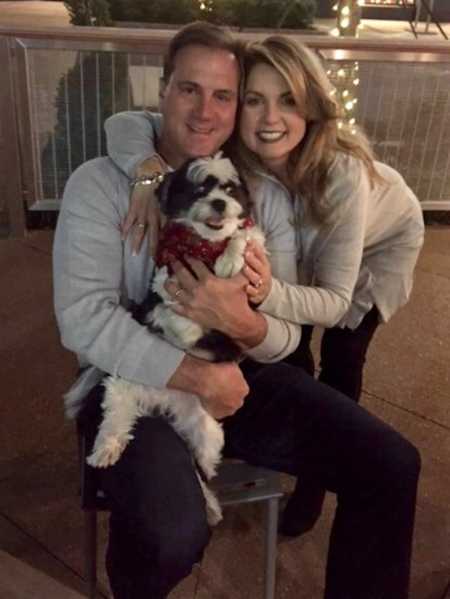 Wife holding arm around husband who is cancer free sitting with dog in his lap