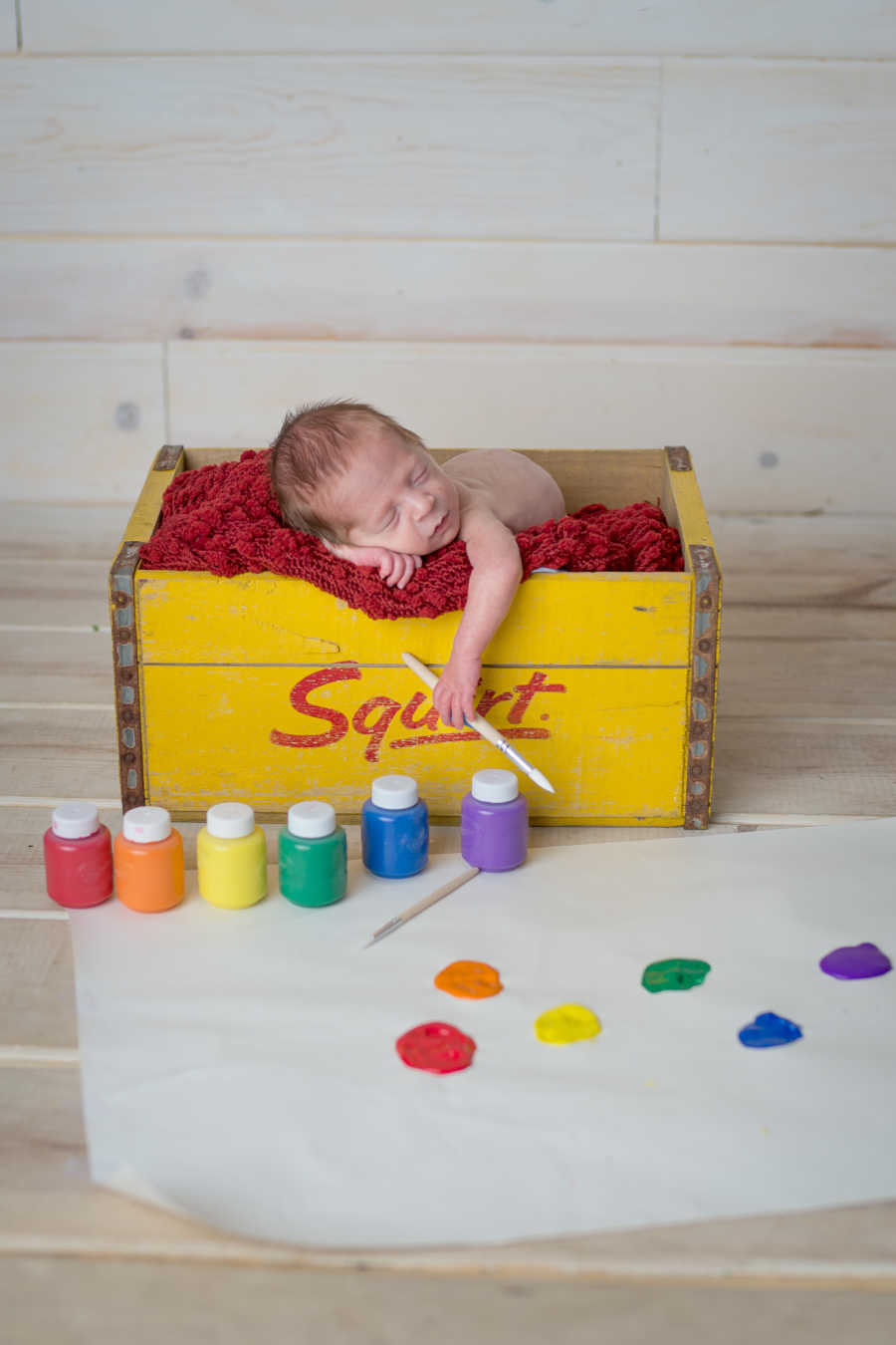 Newborn sitting in wooden box holding paintbrush next to rainbow paint