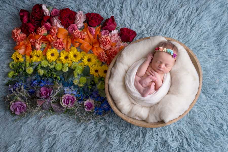 Newborn sitting in basket next to rainbow colored flowers