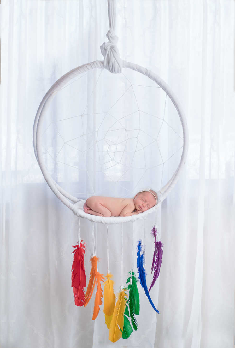 Newborn lying in large dream catcher with rainbow feathers hanging from it