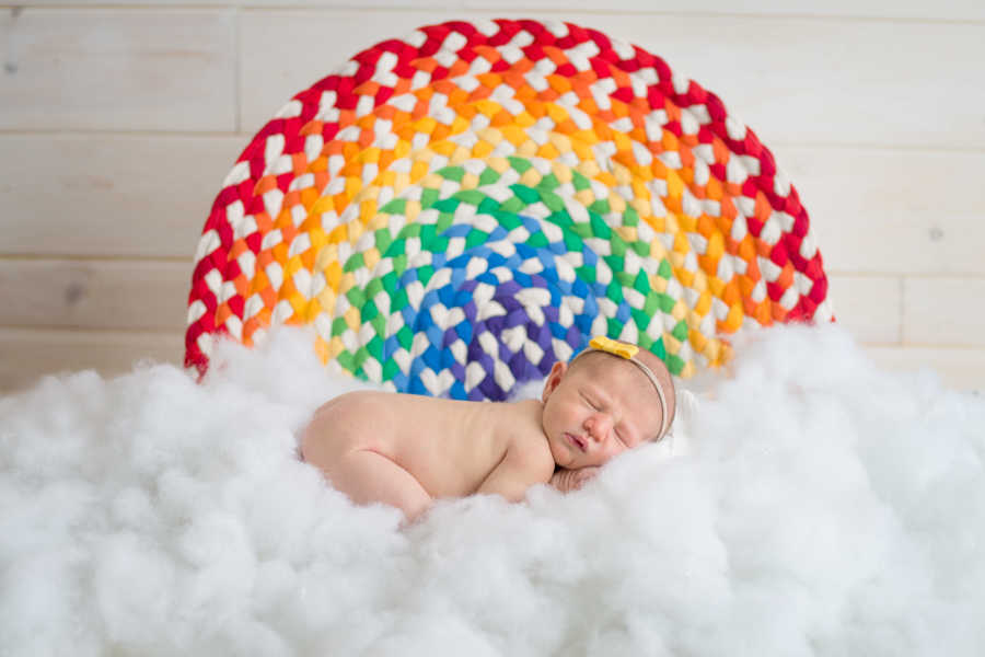 Newborn laying on fluffy white material with rainbow fabric behind 