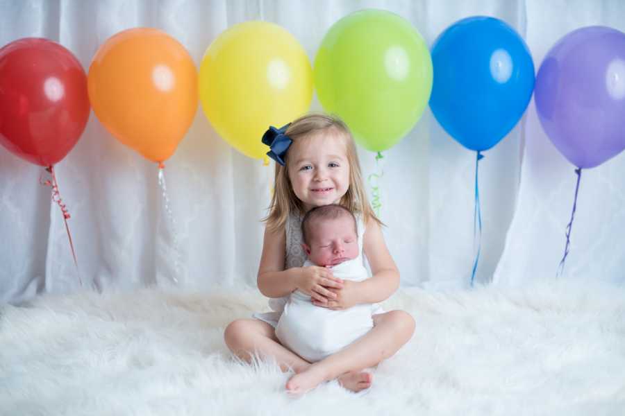 Toddler sitting with newborn sibling in her lap with rainbow balloons behind them