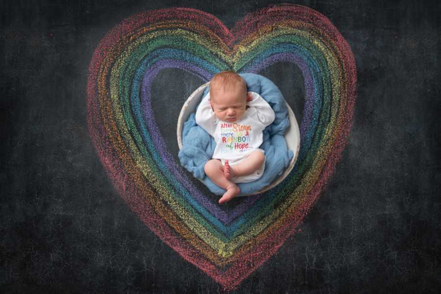 Newborn sits in basket enclosed in chalk drawing of heart in colors of rainbow