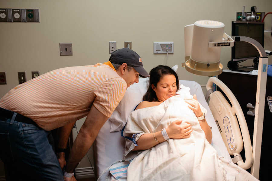 Husband leans over wife who is in hospital bed who is holding adopted newborn 