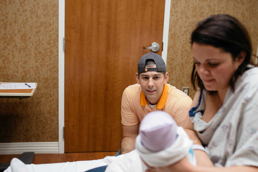 Mother holds adopted newborn up for husband who is sitting in chair beside her