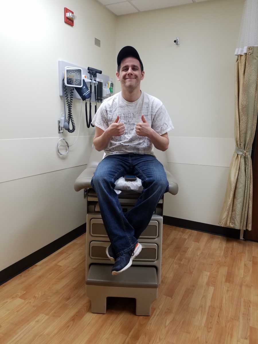 Gay son sitting on doctors table with two thumb up before donating kidney to mother