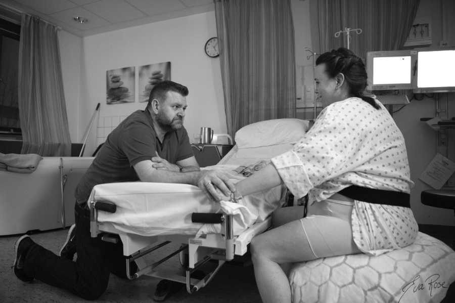 Husband reaches over hospital bed to hold arms of pregnant wife who is sitting on pregnancy ball
