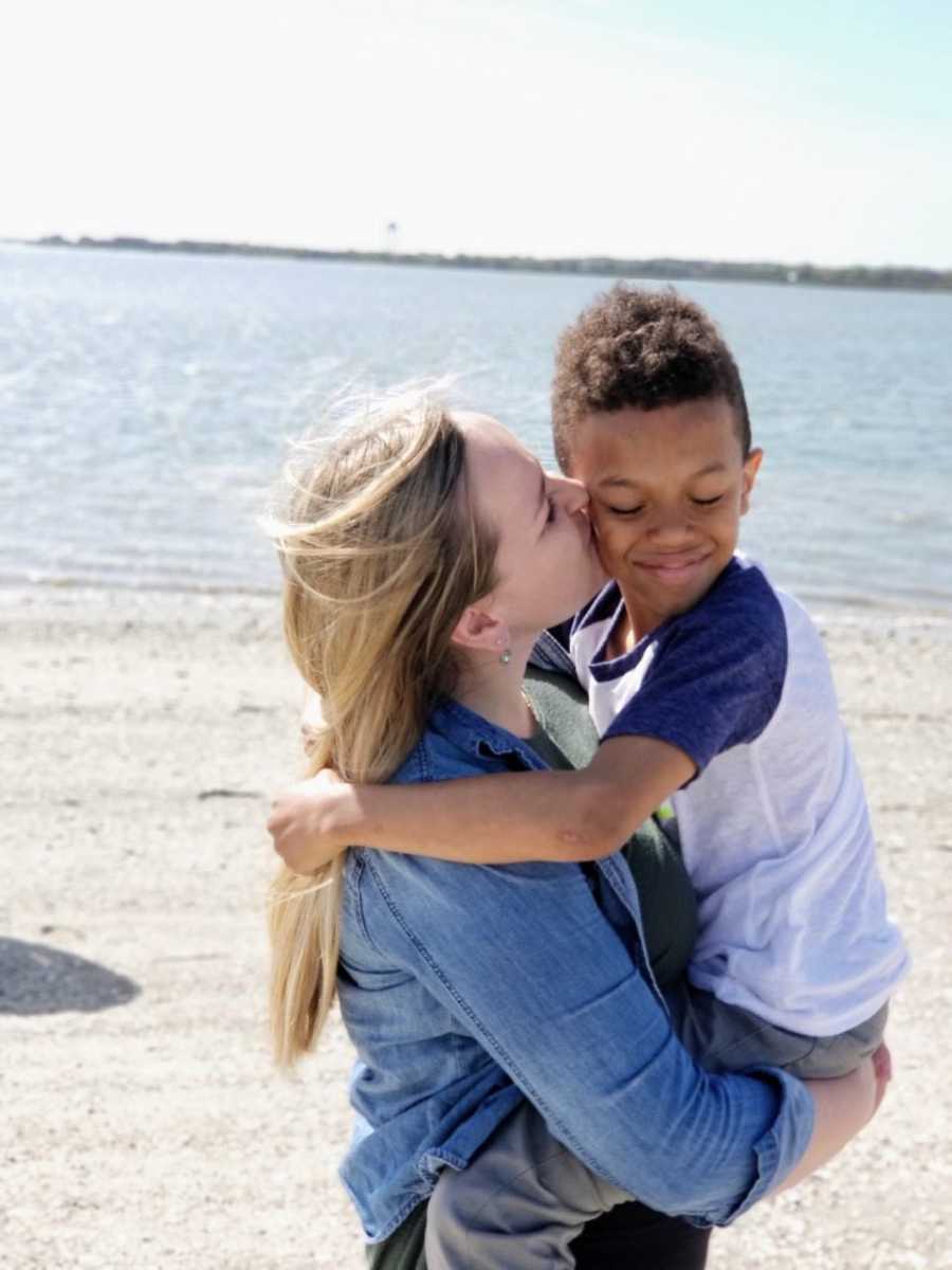 Woman holds adopted son while giving him a kiss on cheek at beach