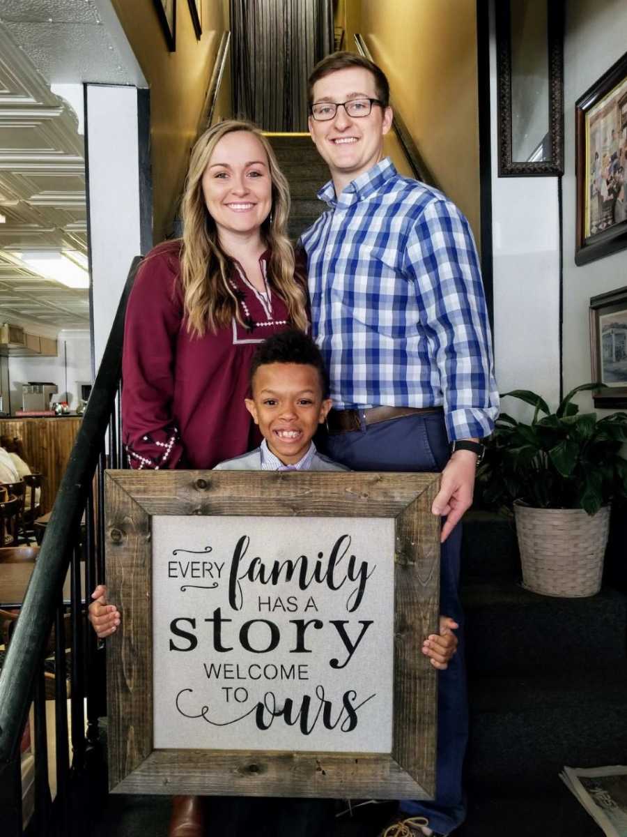 Mother and father stand with adopted son who holds sign saying, "every family has a story welcome to ours"