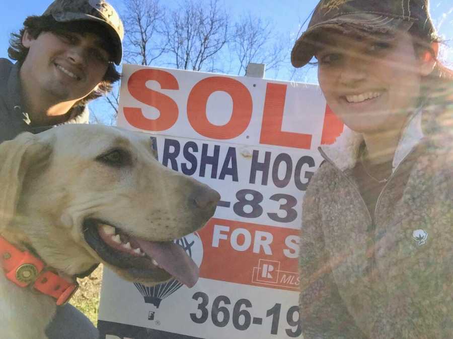 Girlfriend smiles in selfie with dog and boyfriend in front of SOLD sign in yard