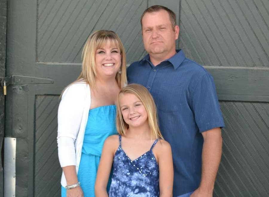 Woman stands smiling beside husband who has since passed from cancer and young daughter