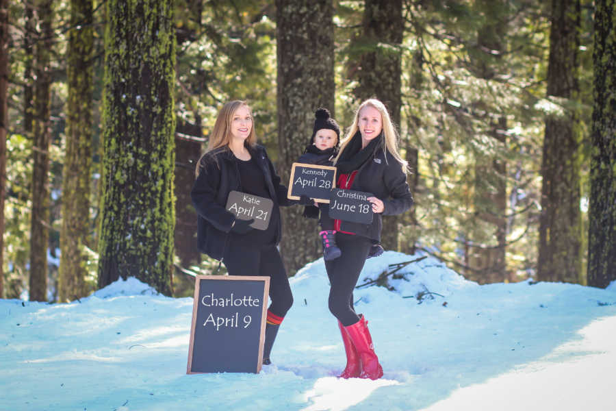 Woman holds firstborn next to wife who are all holding chalk boards with their birthdays and birthday of future child