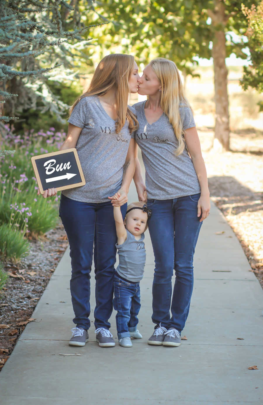 Wives kissing while holding hands with firstborn while pregnant wife through IVF holds sign saying "bun" pointing to stomach