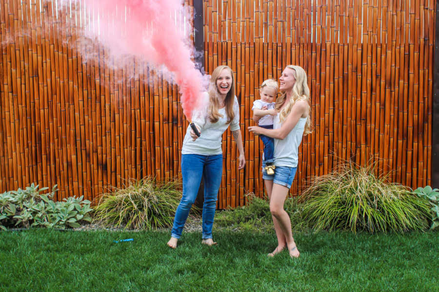 Woman holds firstborn while her wife holds can of pink smoke at gender reveal 