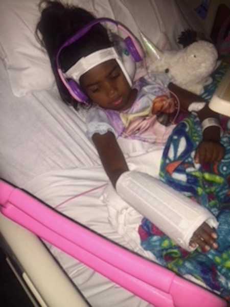 Adopted toddler girl lies in hospital bed with headphones on and case on her arm 