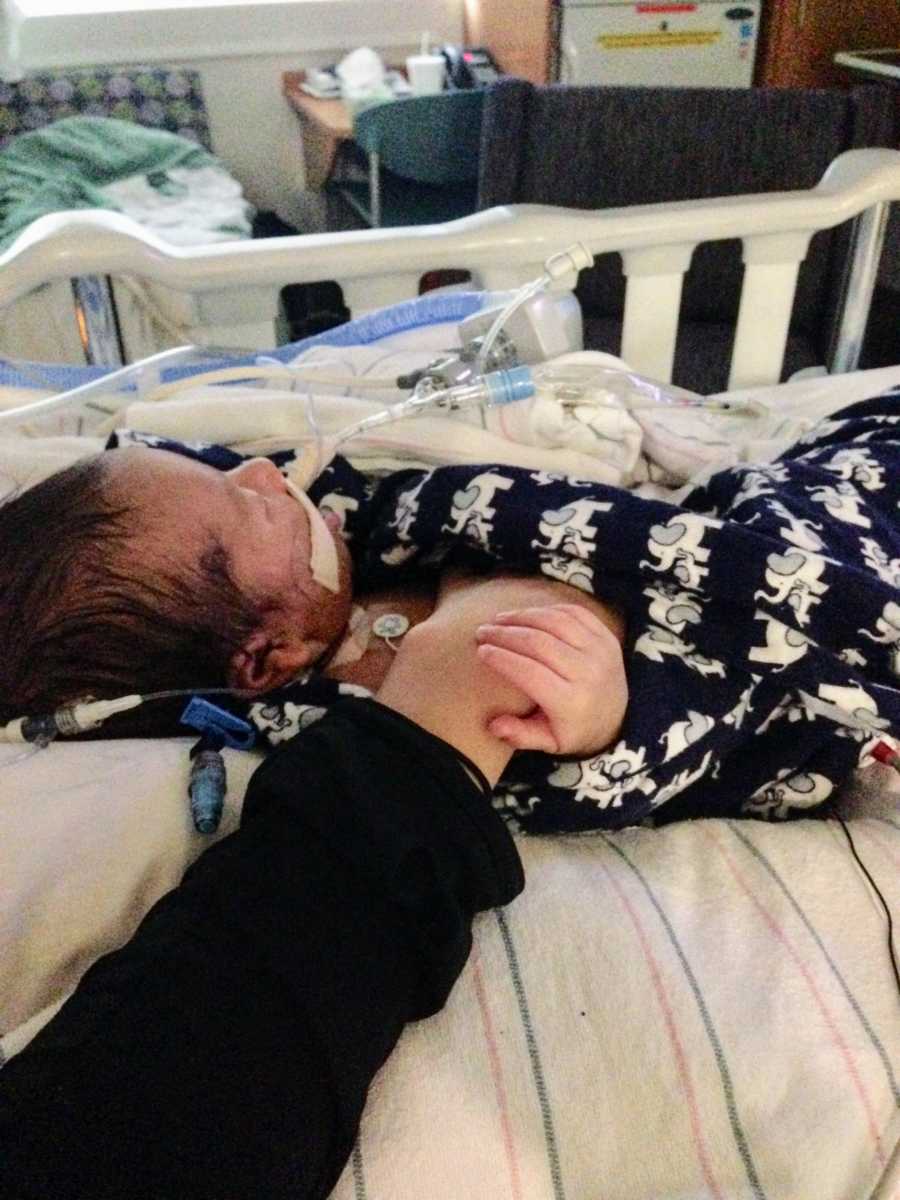 Baby laying in hospital bed hooked up to machines holding mother's hand