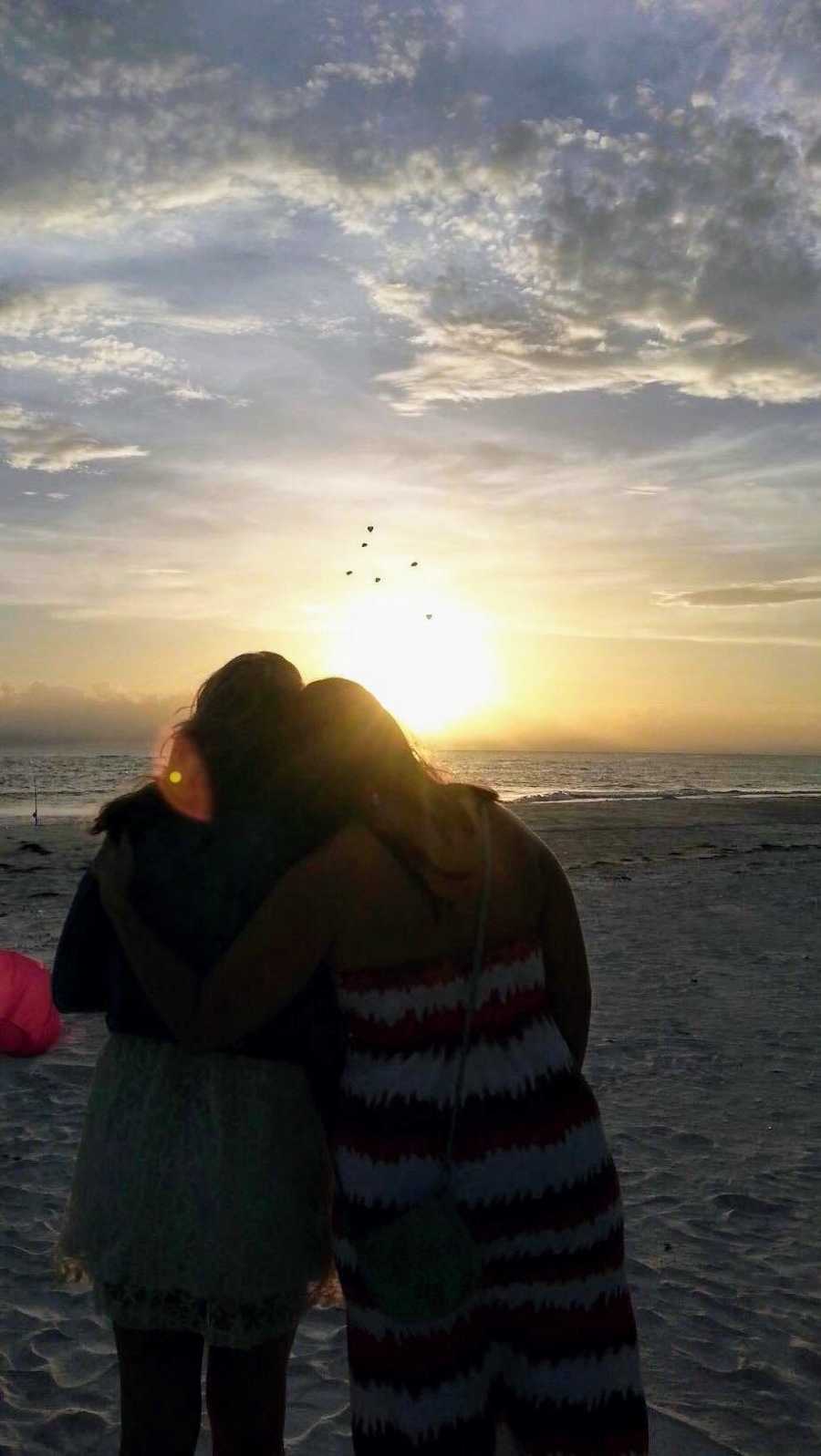 Friends who met on grief journey stand arm in arm looking out at ocean 