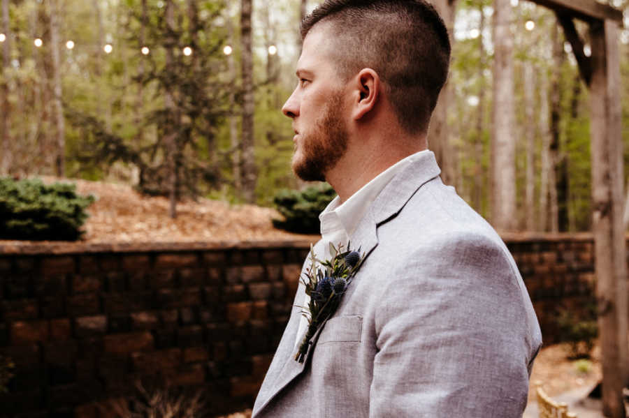 EMT boyfriend stands in gray suit with boutonniere looking out into woods 