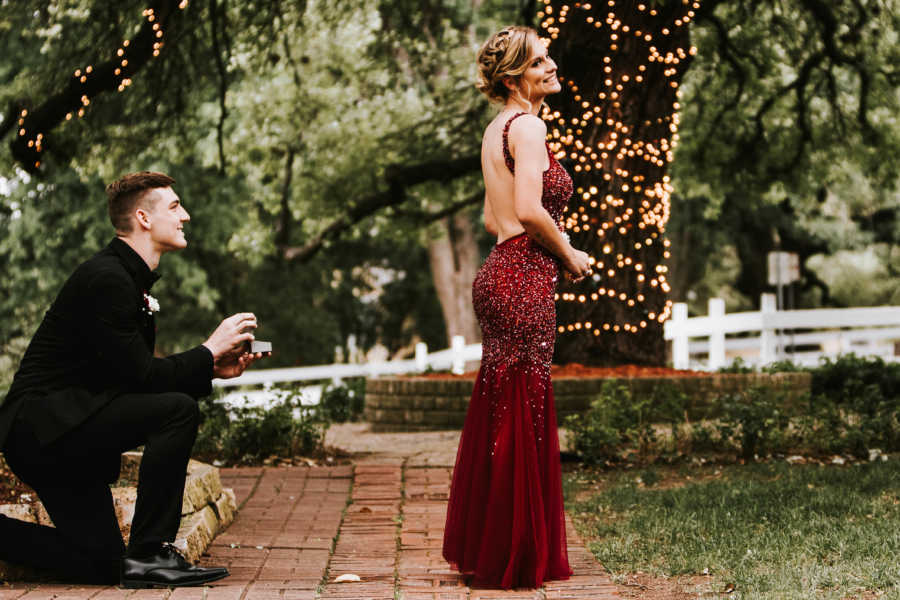 Girlfriend in prom dress stands with back turned to boyfriend who is down on one knee