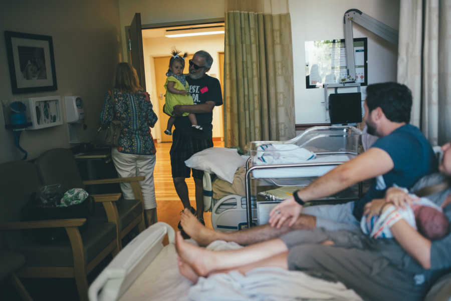 Mother and father lay in hospital bed holding newborn while grandparents bring in firstborn conceived through IVF