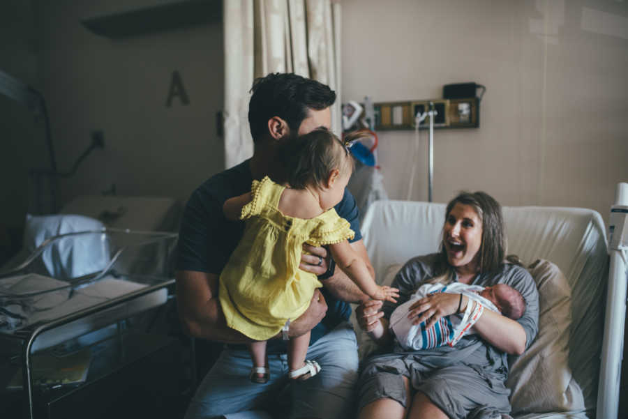 Mother smiles at firstborn in husbands arms while she holds natural born baby