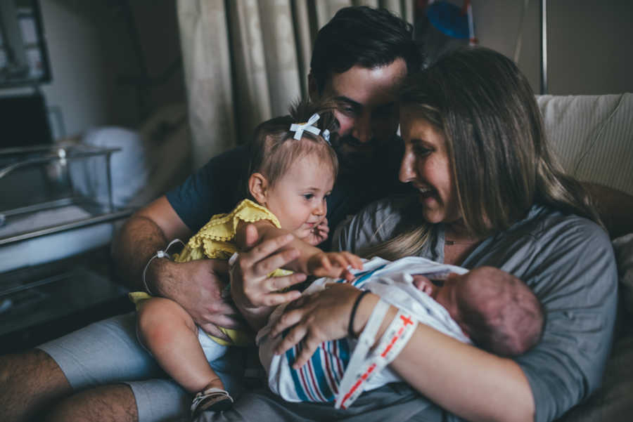 Firstborn conceived through IVF sits in dad's lap reaching for newborn sibling in moms arms
