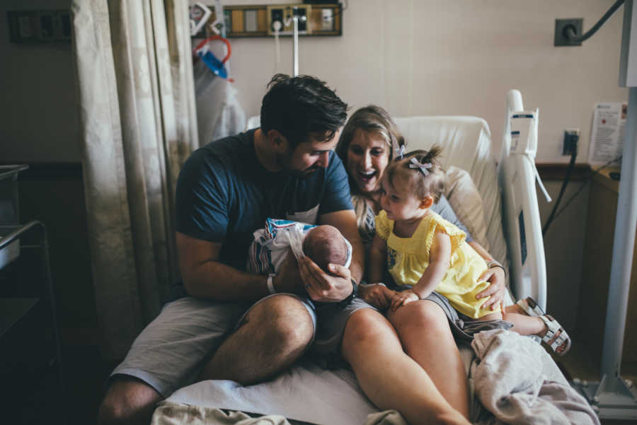 Mother and father sit in hospital bed holding infant newborn and three month old daughter conceived through IVF