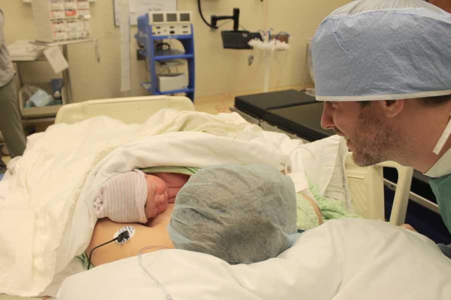 Mother holds newborn to chest after c-section while husband stands behind looking at baby