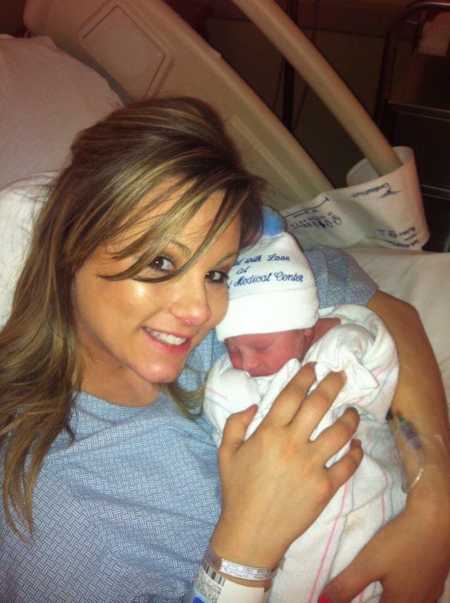 Woman smiling in hospital bed holding newborn swaddled in blanket and wearing hat