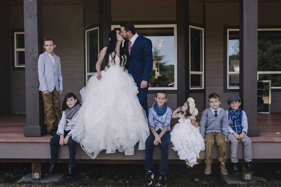 Bride and groom stand kissing on porch with their children sitting on either side of them