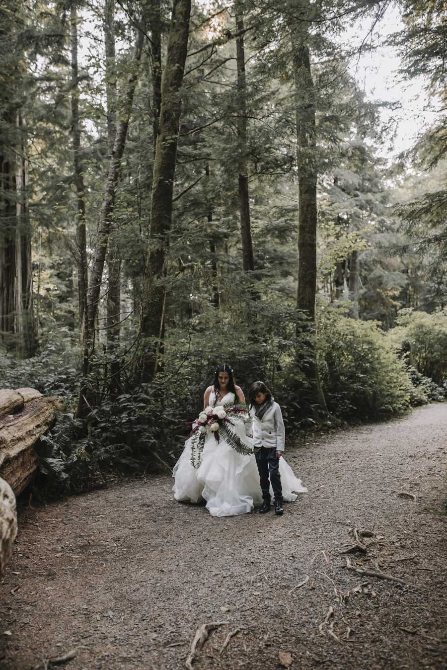 Bride walks through path in forest holding hands with son who is giving her away