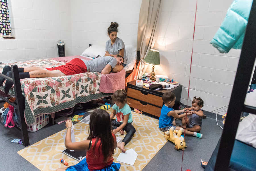 Mother sits on bed with husband laying head on her lap while their four children play on floor of homeless shelter