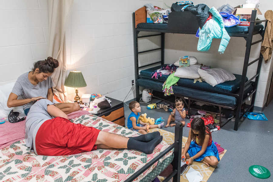 Mother sitting on bed with husband laying in her lap and three kids playing on the floor of homeless shelter